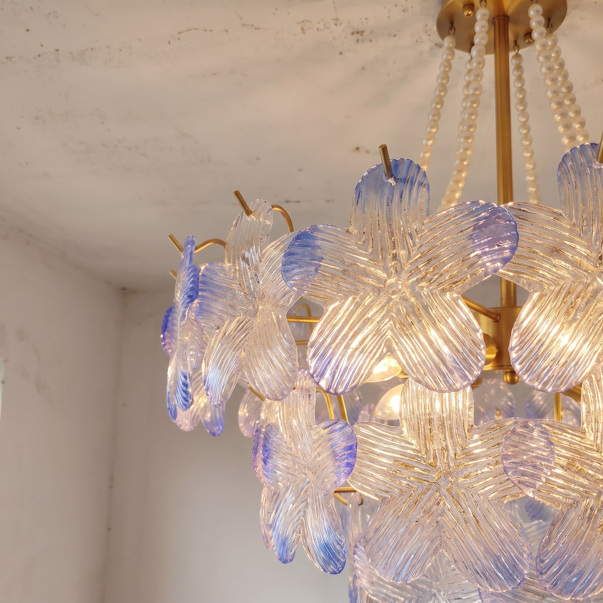 Lustre allumé diffusant une lumière chaude et accueillante, ses 12 ampoules E14 faisant scintiller les nuances pastel bleutées et rosées du verre.