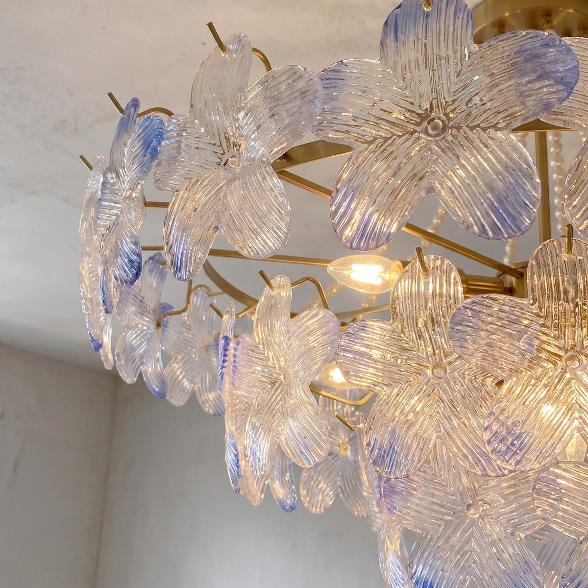 Lustre de plafond allumé, mettant en valeur la lumière douce diffusée par les douze ampoules E14 à travers les pétales de verre irisé bleu-lavande.