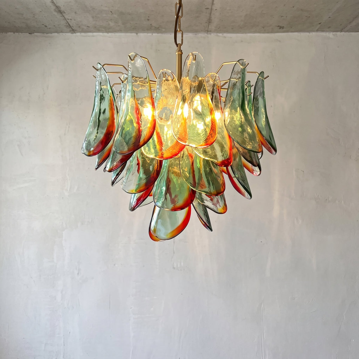Lustre suspension style Murano vénitien, avec des pétales de verre soufflé vert et rouge ambré, éclairant un salon moderne.